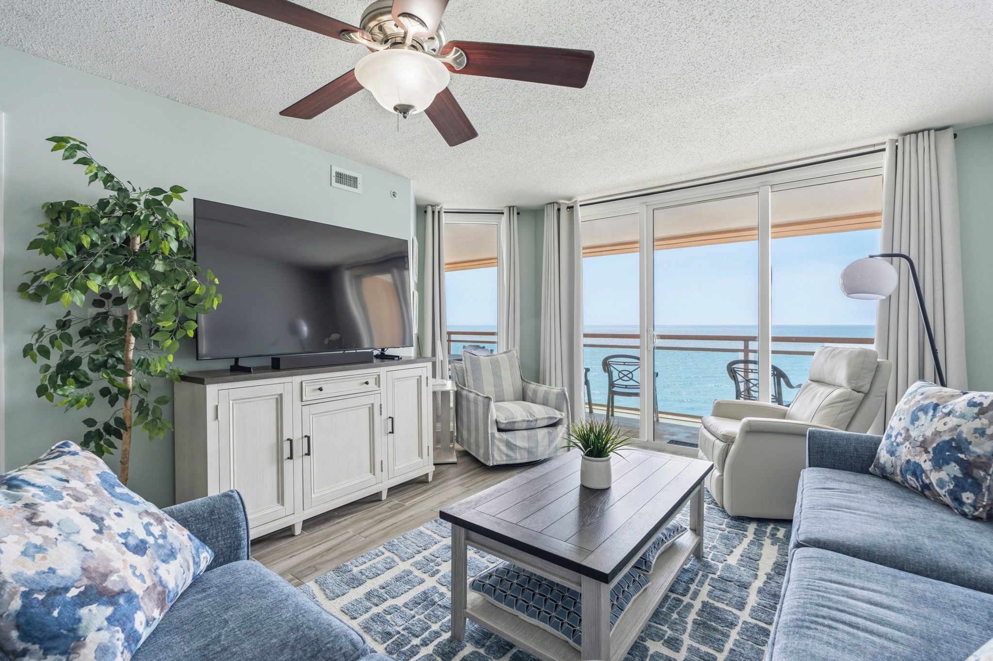 Ein modernes Wohnzimmer mit einem Flachbildfernseher, einem Deckenventilator, einem großen Fenster mit Blick aufs Meer und einem Balkon mit Sitzgelegenheit.