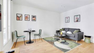 Een moderne woonkamer met een grijze bank, twee stoelen, een klein rond tafeltje en een salontafel. Er hangen ingelijste foto's aan de muur en een afstandsbediening ligt op de vloer.