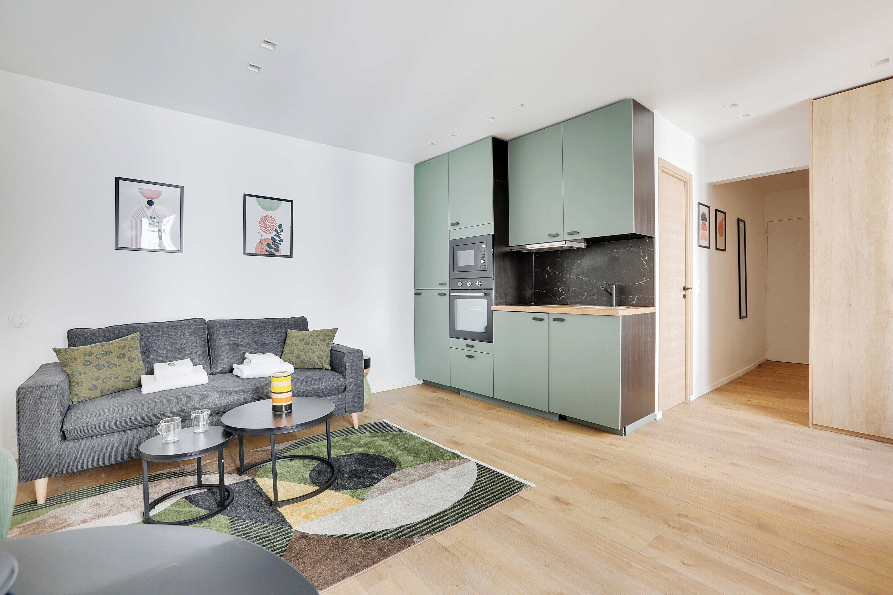 A modern living room with a grey sofa, a small coffee table, and a kitchen with green cabinets and a microwave.