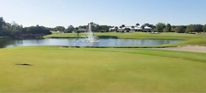 En golfbane med en sø, en springvand og en klubhus i baggrunden.