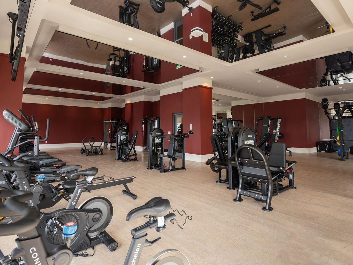 A gym with multiple exercise machines, including treadmills and weight stations, with a high ceiling and red walls.