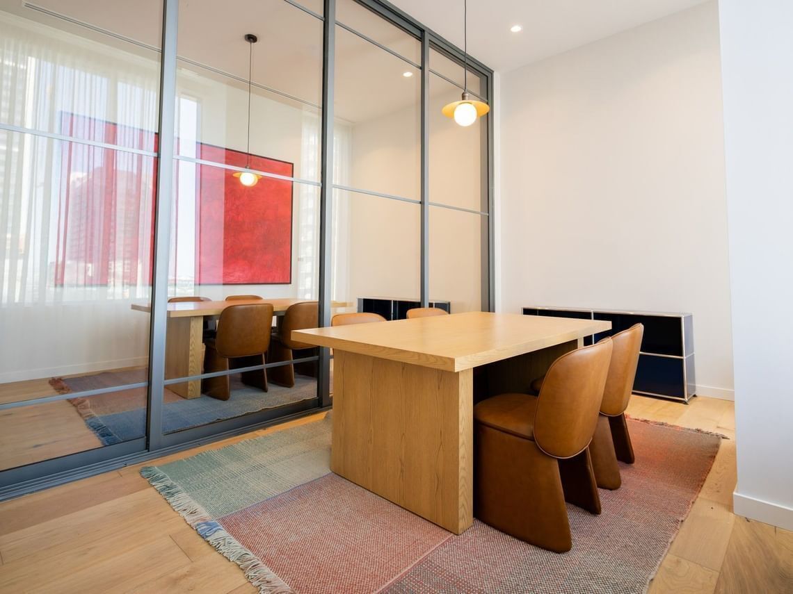 A modern dining area with a wooden table and leather chairs, large windows with a view, and a glass partition.