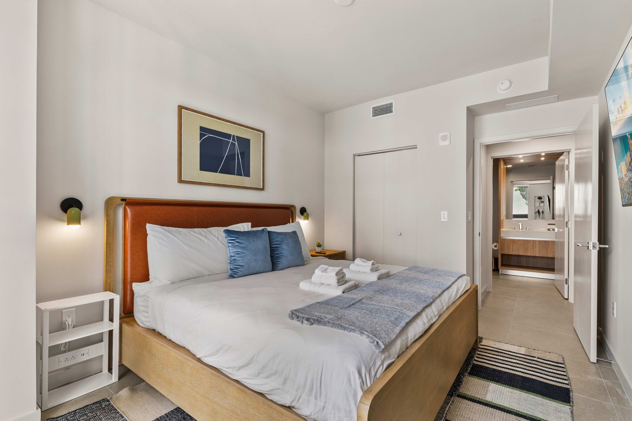 A hotel room with a bed, bedside table, wall-mounted artwork, and a bathroom visible through an open door.