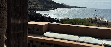 A beachside patio with a lounge chair, a thatched roof structure, and a view of the sea and distant hills.