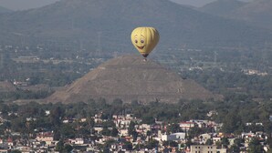 A hot air balloon with a smiling face, flying over a town with buildings and hills in the background. - Complejo Tur ístico Mixcóatl  (San Martín de las Pirámides)