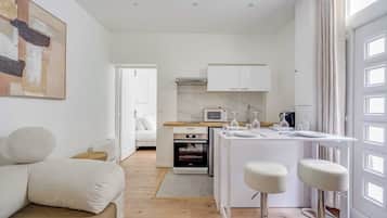 Una cocina moderna con armarios blancos, microondas integrado y horno pequeño. Hay una zona de comedor con una mesa blanca y dos taburetes altos.