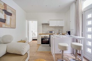 A modern kitchen with white cabinetry, a built-in microwave, and a small oven. There is a dining area with a white table and two bar stools. - Modern Apartment 1br/4p - Levallois (Levallois-Perret)