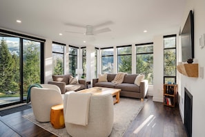 A modern living room with large windows, a ceiling fan, and a fireplace. - Cedar Sky at Ravencrest (Whistler)