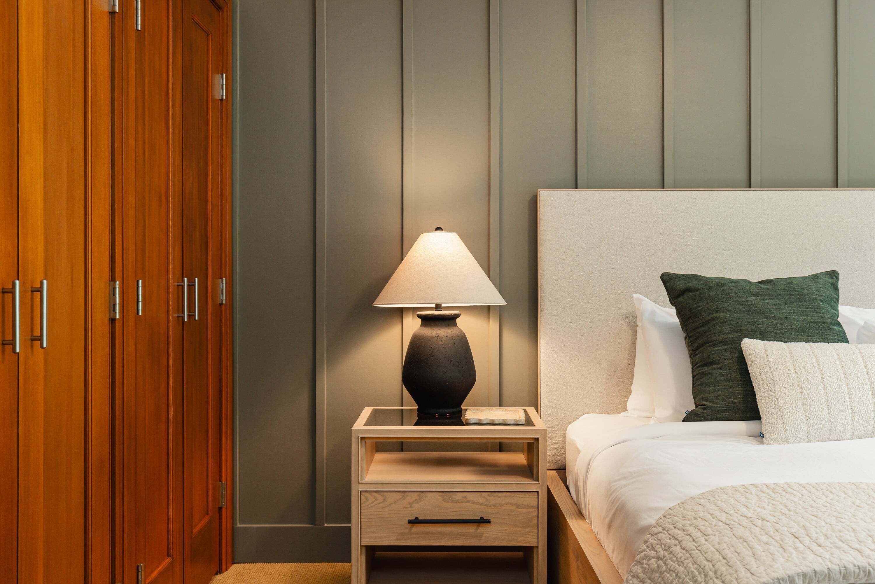 A hotel room with a wooden wardrobe, a bedside table with a lamp, a bed with white bedding and a green pillow, and a grey wall.