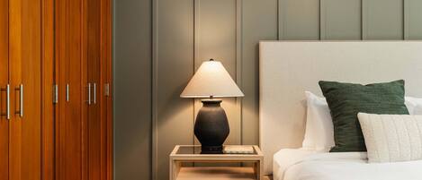 A hotel room with a wooden wardrobe, a bedside table with a lamp, a bed with white bedding and a green pillow, and a grey wall.