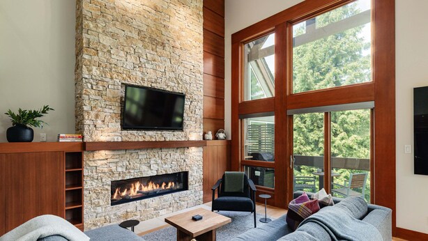 A living room with a stone fireplace, a flat-screen TV, a sofa, and a view of the outdoors through large windows. - Northlight Retreat at Alpine Greens (Whistler)