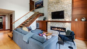 A modern living room with a stone fireplace, a large sofa, a wooden staircase, and a flat-screen TV mounted on the wall. - Northlight Retreat at Alpine Greens (Whistler)