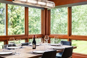 Een gedekte eettafel voor zes personen, met wijnglazen, borden en een fles wijn, voor grote ramen met uitzicht op groen.