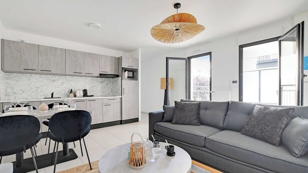 Un salon moderne avec un canapé gris, des fauteuils noirs, une table basse ronde et une cuisine en arrière-plan.