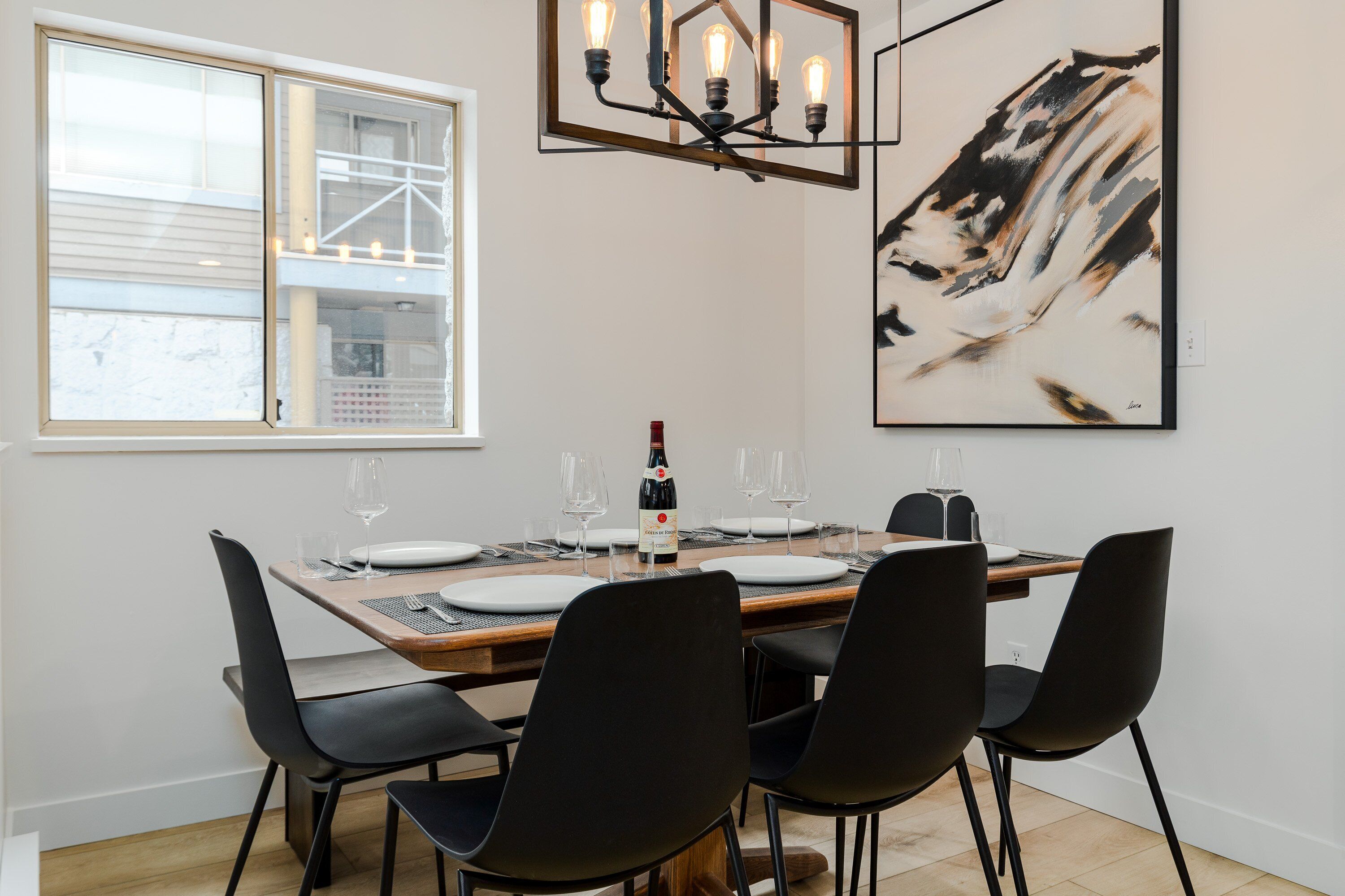 A dining area with a wooden table set for four, black chairs, a chandelier, and a large abstract painting on the wall.