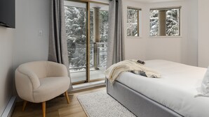 A bedroom with a bed, a chair, a large window with a view of snow-covered trees, and a balcony. - The Altitude at Valhalla (Whistler)