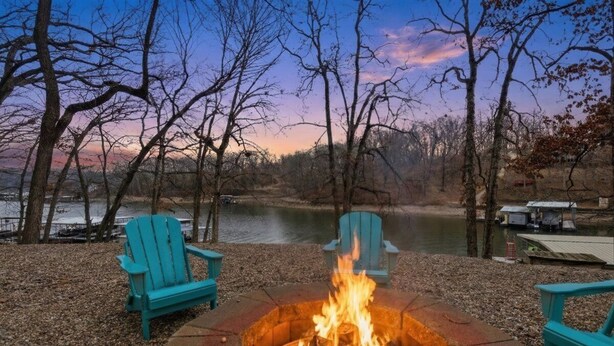 Un área al aire libre junto al lago con una fogata, dos sillas Adirondack turquesas y vistas al lago y los árboles.