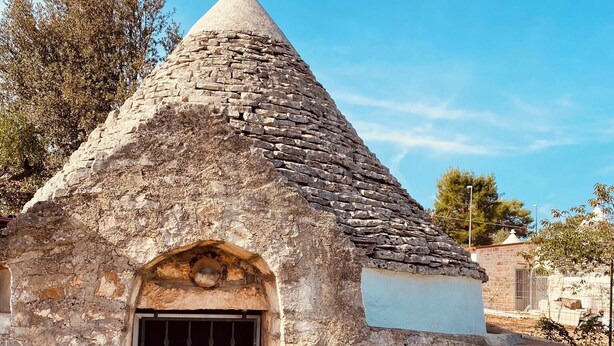 Un bâtiment en pierre surmonté d’un toit conique, une fenêtre avec des barreaux en métal et une bouteille verte.