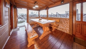 Une terrasse en bois avec une table de pique-nique et des bancs, un mur en pierre et de grandes fenêtres donnant sur un paysage.