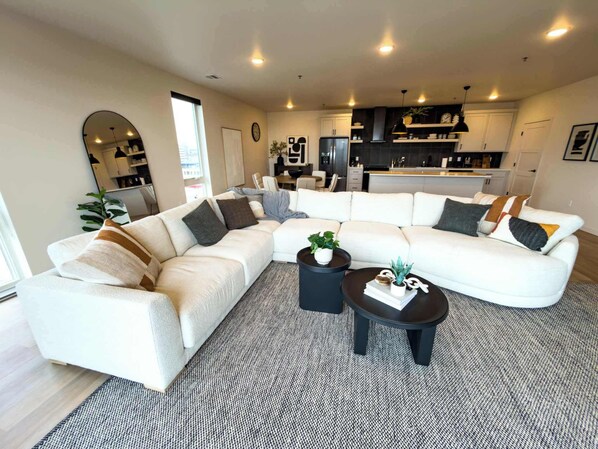 A modern living room with a sectional sofa, a coffee table, and a kitchen area in the background.