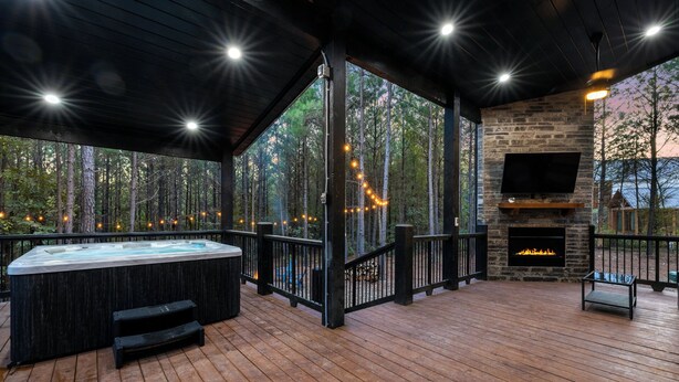 A covered outdoor deck with a hot tub, stone fireplace, and a TV mounted on the wall. - Timbuktu with You: Hot Tub, Foosball, WiFi, Pet Friendly (Broken Bow)