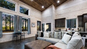 A spacious living room with a large couch, ottoman, and a dining area with a table and chairs. There are large windows offering a view of the forest. - Timbuktu with You: Hot Tub, Foosball, WiFi, Pet Friendly (Broken Bow)