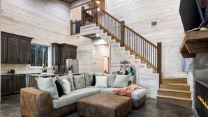 A spacious living area with a wooden staircase, a large sectional sofa, and a kitchen with dark cabinets. - Timbuktu with You: Hot Tub, Foosball, WiFi, Pet Friendly (Broken Bow)