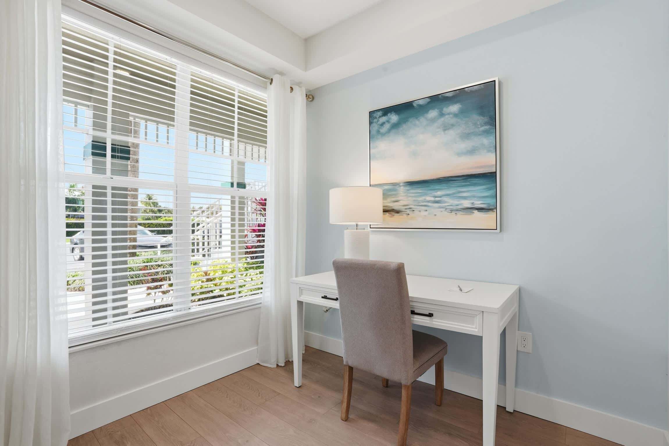Una habitación con escritorio, silla, ventana con persianas y un cuadro de una playa.