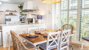 Une cuisine moderne avec des meubles blancs, un îlot central et un coin repas agrémenté de chaises en osier et d’une table en bois.