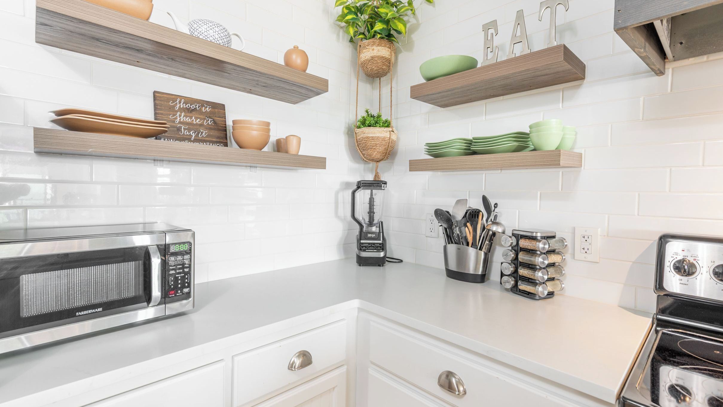 A kitchen with white cabinets, a microwave, a blender, and shelves with dishes and plants.