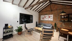 A loft-style living space with exposed wooden beams, a brick wall, a grey sofa, a wooden dining table, and a coffee machine. - Apartamento Hija Mía (Medellín)