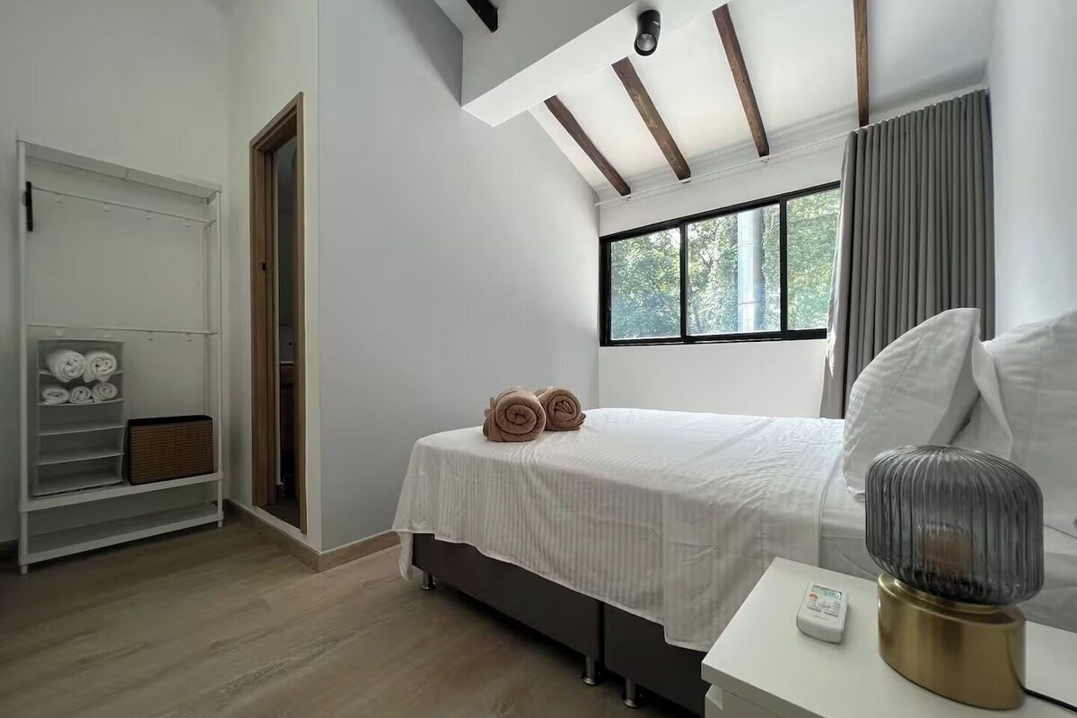 A bedroom with a bed, a bedside table with a lamp, a window with curtains, and a wardrobe with towels.