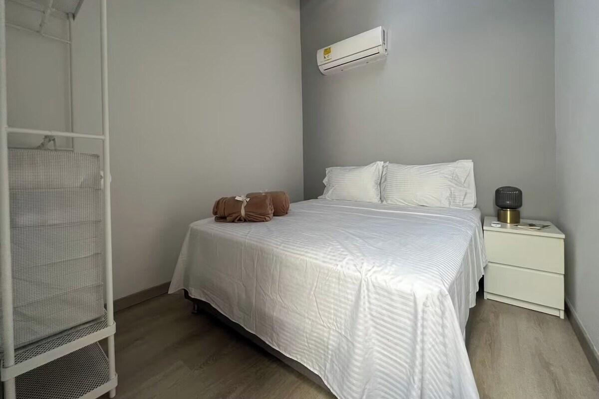 A bedroom with a bed, white bedding, a nightstand, an air conditioning unit, and a metal shelf unit.