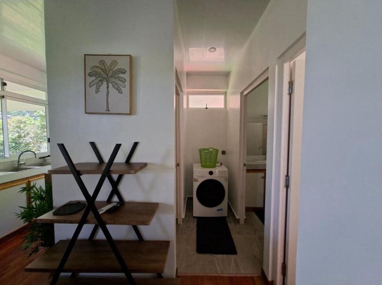 A hallway with a washing machine, a green basket, and a palm tree artwork on the wall.