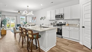 A modern kitchen with white cabinets, a large island, bar stools, and a dining area with a view. - Cozy Escape Retreat – Luxury Brand New 3BR/3BA (Myrtle Beach)