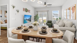 A modern living room with a dining table set for four, a sofa, a ceiling fan, and a large window. - Cozy Escape Retreat – Luxury Brand New 3BR/3BA (Myrtle Beach)