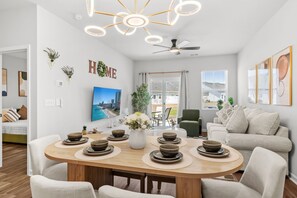 A modern living room with a dining table set for four, a sofa, a ceiling fan, and a large window. - Cozy Escape Retreat – Luxury Brand New 3BR/3BA (Myrtle Beach)
