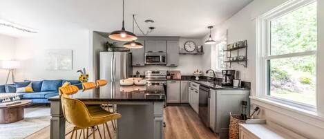 Una cocina moderna con una isla central, taburetes de bar, iluminación colgante y vistas al exterior.