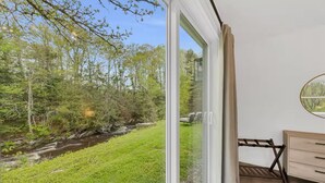 Una habitación con puerta corrediza de vidrio que da a un espacio exterior con césped, árboles y un arroyo.