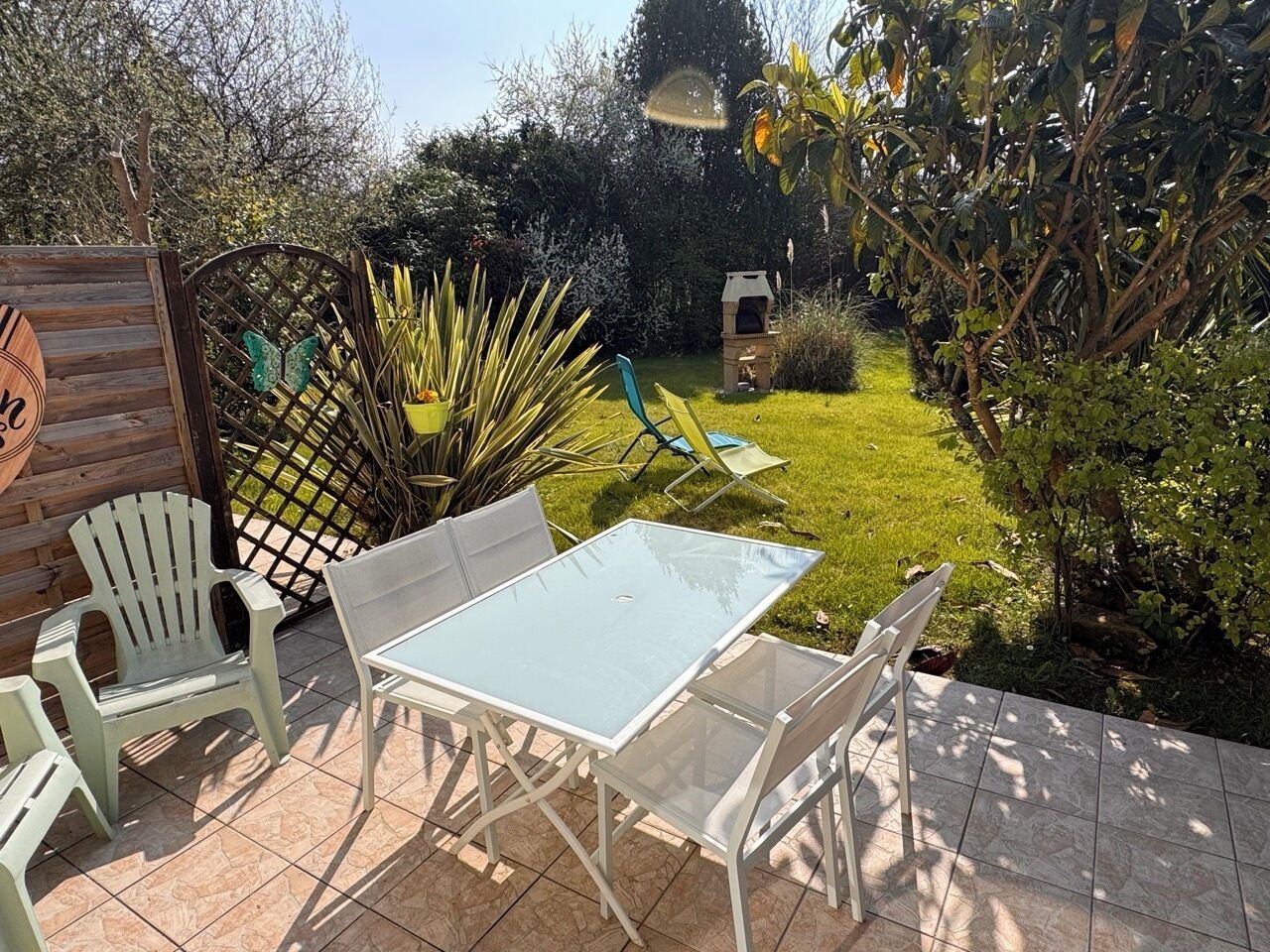 A patio with a glass table and white chairs, surrounded by greenery and a wooden fence.