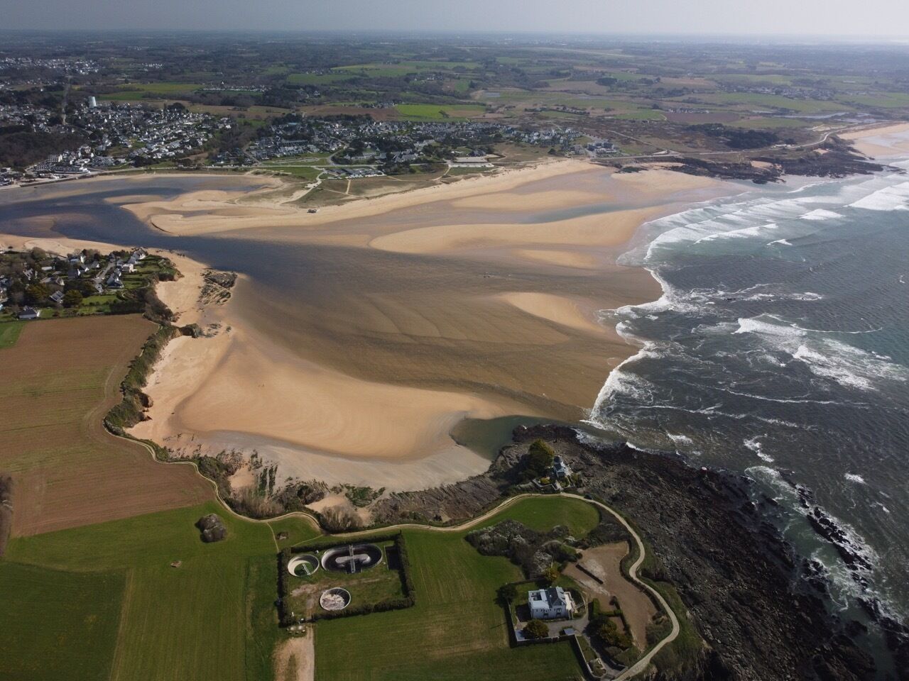 Vista aérea de uma área costeira com uma praia de areia, um pequeno povoado e um trecho de costa rochosa.