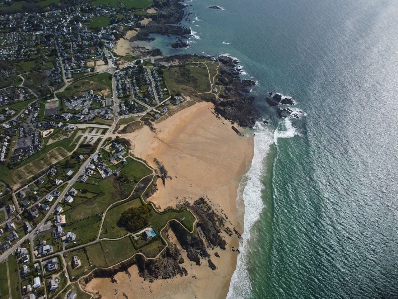 An aerial view of a coastal area with residential buildings, a sandy beach, and rocky coastline.