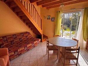 A cozy room with a wooden staircase, a patterned sofa, a round table with chairs, and a view through a large window. - Le pouldu plein sud - Large Garden - Close to beaches (Clohars-Carnoët)