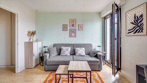 A modern living room with a grey sofa, two side tables, a wooden coffee table, and a large window with a view. - Spacious Studio 2P - Near La Defense (Courbevoie)