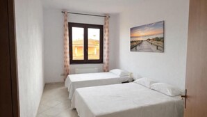 Una habitación con dos camas, una ventana con cortinas, un cuadro en la pared y piso de baldosas.