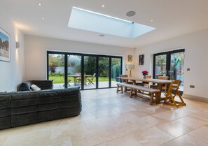 A spacious living area with a sofa, dining table, and benches, large sliding glass doors to a garden, and a skylight. - Large 6BR House Near Goodwood Garden Parking (Chichester)