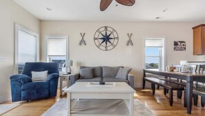 A living room with a sofa, a coffee table, a blue armchair, a dining table with benches, and a kitchen area. - Coastal Cabana, an island escape (Carolina Beach)