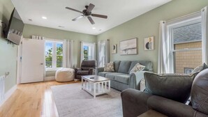 A spacious living room with a ceiling fan, a grey sofa, a coffee table, and a large window with curtains. - Coastal Cabana, an island escape (Carolina Beach)