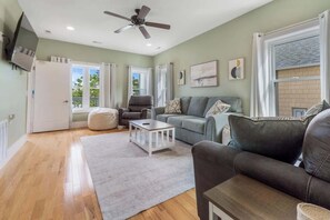 Ein geräumiges Wohnzimmer mit Deckenventilator, einem grauen Sofa, einem Couchtisch und einem großen Fenster mit Vorhängen.