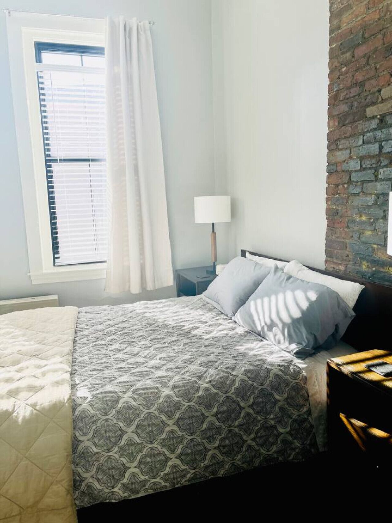 Un dormitorio con cama, mesita de noche, lámpara, ventana con persianas y pared de ladrillo.